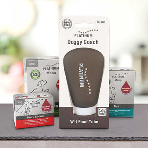 PLATINUM MENU Mini mokra hrana in DoggyCoach - nagrada za tvojega psa: Pripomoček: DoggyCoach tuba (89 ml) stoji pokončno pred tremi škatlami mokre hrane s tremi okusi v ozadju.
