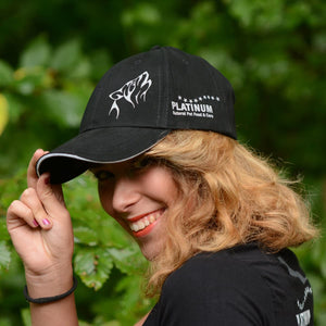 Nasmejana oseba z valovitimi svetlimi lasmi nosi kapo Ekskluzivna PLATINUM kapa s šiltom-tvoja nagrada za zvestobo!-izdelek PLATINUM z grafiko volka in besedilom, ki stoji na prostem med zelenim listjem.