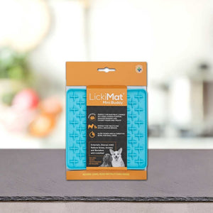 Podloga LickiMat® - BUDDY by PLATINUM v modri barvi z oranžno embalažo stoji pokonci na pultu, idealna za poticanje sporog hranjenja psa, na zamegljenem kuhinjskem ozadju.