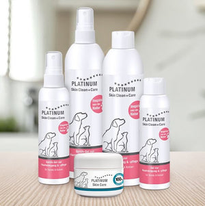 Na svetli površini z zamegljenim kopalniškim ozadjem je razporejenih pet izdelkov Platinum Skin Clean+Care za nego kože hišnih ljubljenčkov, vključno s stekleničkami z razpršilom in majhnim kozarcem. Na etiketah izdelkov sta prikazani silhueti psa in mačke.