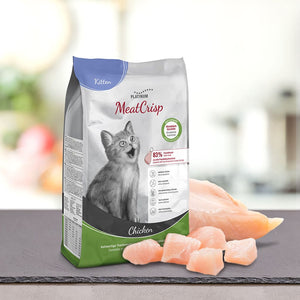 Vrečka PLATINUM MeatCrisp Kitten Chicken, Suha Hrana za Mačke leži na pultu ob kosih surovega piščanca. Njegova embalaža s sivo mačico in podrobnostmi Freshmeatdryer izstopa v nežno zamegljenem ozadju.